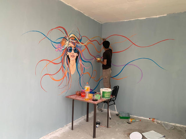 Person painting a colorful mural of a woman's face on a wall with art supplies on a table.