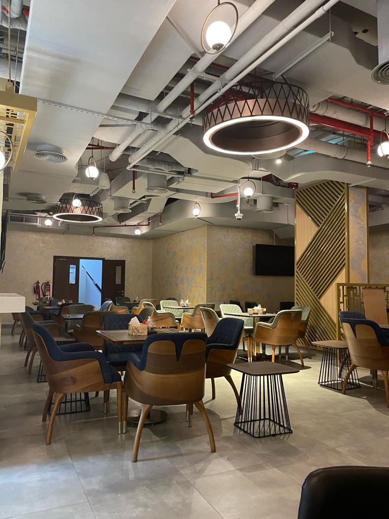 A modern indoor cafe or lounge space with multiple dining tables, upholstered chairs in brown and blue, exposed ceiling ducts and industrial pendant lights, and light stone tile flooring.