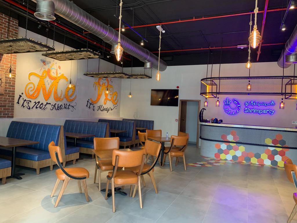 A modern cafe interior with blue upholstered booth seating, orange-brown chairs, hanging pendant lights, exposed ceiling ducts, and a reception desk with colorful hexagon decals. Wall art and signage are visible, including a large orange 'Me' graphic and a blue logo on the counter.