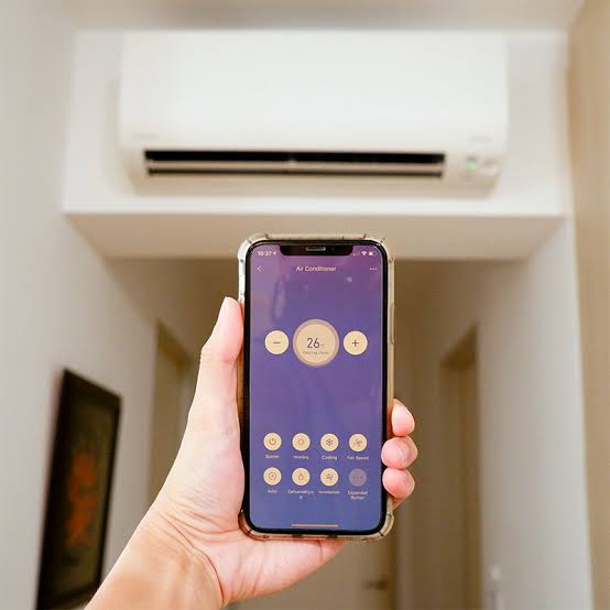 A hand holds a smartphone showing a smart air conditioner control app screen in front of a real indoor home environment with a wall-mounted air conditioner visible in the background.